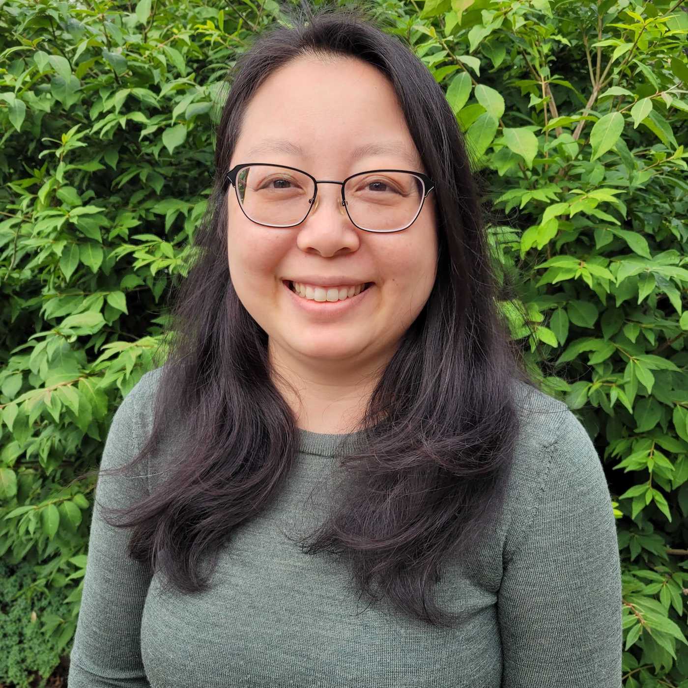 Headshot of author Phuong Truong, smiling, wearing eyeglasses and with long dark hair against green leafy background.